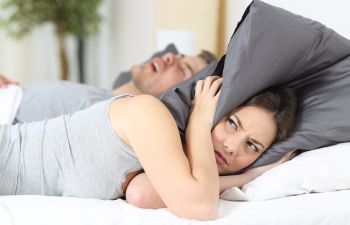 A snoring man sleeping next to a fed-up woman covering her ears with a pillow.