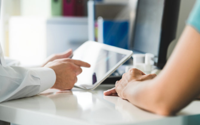 A doctor reviewing digital records on a tablet while talking to a patient.