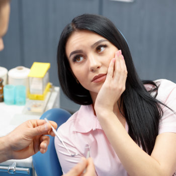black-haired woman with a dental issue rubbing her cheek while discussing the problem with the dentist