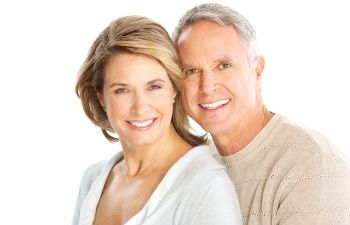 A smiling older couple poses closely against a white background. The woman has shoulder-length blonde hair, and the man has short gray hair. Both are wearing light-colored clothing.