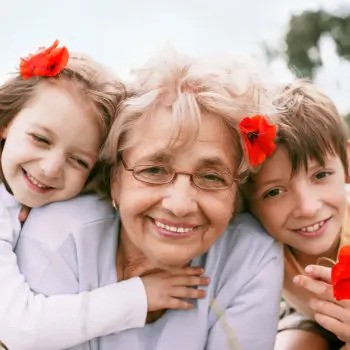 An elderly woman with gray hair and glasses smiles while a young girl and boy, each with a red flower in their hair, embrace her from both sides.