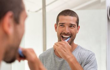 A man brushing his teeth in front of a bathroom mirror, wearing a gray t-shirt.