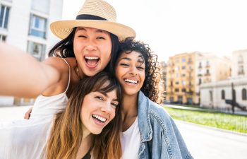 Three young women smiling and taking a selfie outdoors in a sunny urban setting with buildings in the background.