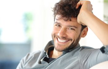 A man with curly brown hair and a beard is smiling at the camera, wearing a gray shirt and sitting indoors with his hand resting on his head.
