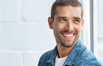 A handsome man with short hair, blue eyes, and a beard smiles broadly, showing his white teeth as he looks off to the side. He is wearing a blue denim shirt against a bright, out-of-focus white background.