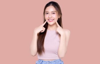 A young woman with long dark hair, wearing a light pink ribbed tank top, smiles broadly and points to her teeth with both index fingers. The background is solid dusty rose/pink.