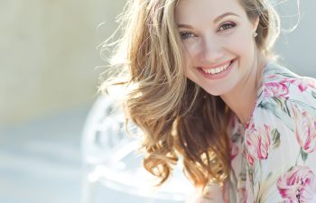A smiling young woman with wavy blonde hair wearing a floral blouse, sitting outdoors in soft natural light.