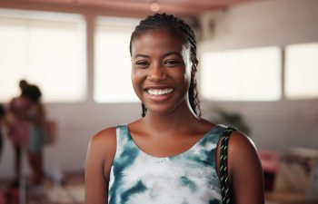 A young woman with braided hair smiles at the camera in a brightly lit indoor setting, with people and furniture blurred in the background.