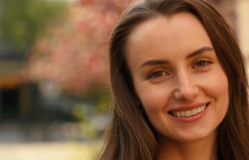 A close-up portrait of a smiling woman with long brown hair and light-colored eyes, looking at the camera against a blurred outdoor background.